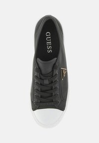 Zapatilla baja negra con punta de goma blanca, cordones negros, logotipo triangular dorado en el costado y la marca "GUESS" en el interior y en la lengüeta.