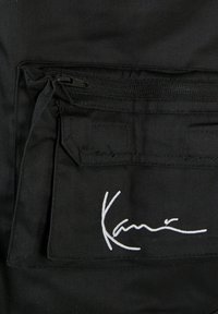 Černá látková taška s přední kapsou na zip. Má bílý vyšitý nápis "Kani" a texturovaný povrch. Minimalistický design.