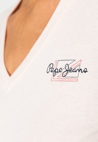 Tričko s V-výstřihem a krátkým rukávem, světle růžové barvy, vyrobené z měkké bavlny, s černým a červeným nápisem "Pepe Jeans" v kurzívě a geometrickými akcenty.