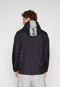 Black windbreaker jacket with a grey hood featuring a logo print. The fabric has a smooth texture, with elastic cuffs and a relaxed fit.