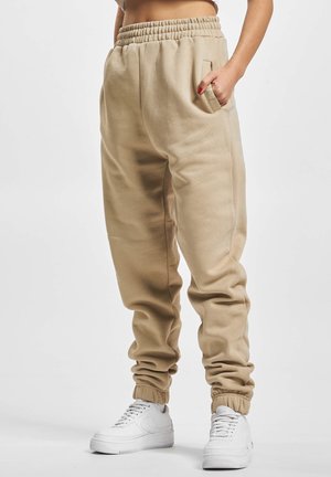 Personne portant un pantalon de jogging beige avec taille et poignets élastiques, main dans la poche, et baskets blanches sur un fond clair uni.