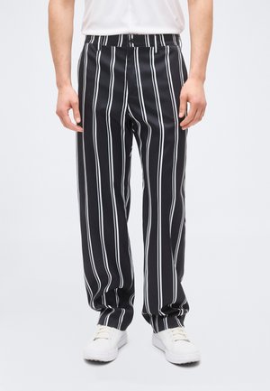 Man die zwarte broek met witte verticale strepen en witte sneakers draagt, staand tegen een effen lichte achtergrond.