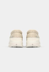 Beige polstrede sneakers med en tykk hvit såle, avrundet bak, og teksturert hæl, med subtile merkevareingrep på siden.