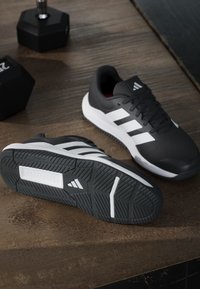 Paire de chaussures de sport Adidas noires et blanches sur un sol en bois à côté de deux haltères hexagonales noires.