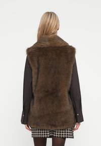 Blonde vrouw die een bruine nepbontvest draagt over een zwart longsleeve shirt en een zwart-witte geblokte rok, met haar rug naar de kijker gekeerd.