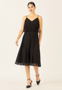 Schwarzes, plissiertes Midikleid mit verstellbaren Spaghettiträgern, schichtigem Oberteil und transparenter Saum, ergänzt durch schwarze spitze High Heels.