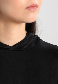 Felpa nera con cappuccio in tessuto texture, caratterizzata da un colletto leggermente plissettato e una vestibilità rilassata, che sottolinea un design minimalista.