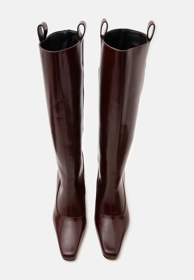 Paire de bottes hautes en cuir marron foncé avec des bouts carrés et des languettes en haut, présentées debout sur un fond clair.