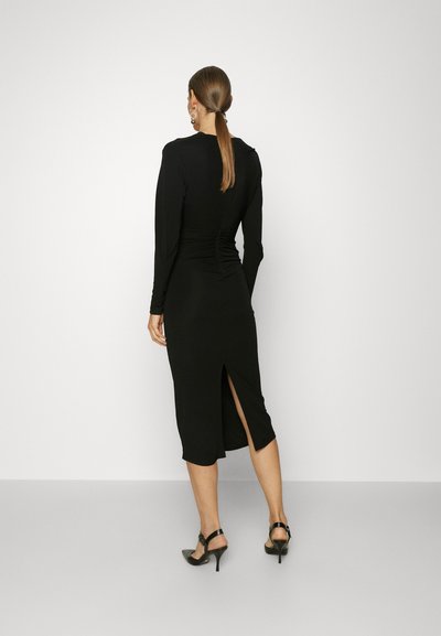 Μαύρο φόρεμα με μακριά μανίκια και εφαρμοστή σιλουέτα, με πτυχώσεις στην πλάτη και σκίσιμο στο πλάι μέχρι το μηρό. Φορεμένο με μαύρες σαμπάνιες με τακούνι.
