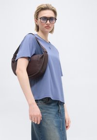 Νεαρό άτομο που φορά γυαλιά ηλίου, ανοιχτό μπλε μπλουζάκι, τζιν φούστα, κρατώντας σκούρο καφέ δερμάτινη τσάντα ώμου σε απλό φόντο.