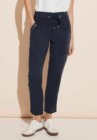 Námořnické joggery se stahovací šňůrkou a kapsami na zip, zkrácená délka a jemná textura. Stylizováno s bílými sportovními teniskami.