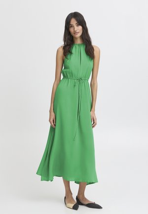 Femme aux longs cheveux foncés portant une robe midi verte sans manches avec une ceinture resserrée à la taille et des ballerines pointues noir et beige, debout sur fond blanc.