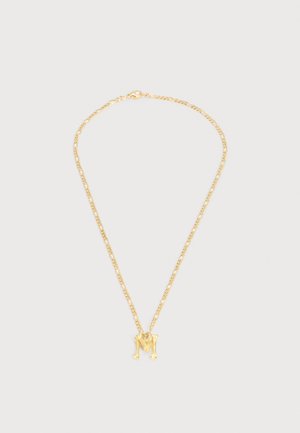 Auksinė grandinėlė su pakabuku, formos raidės „M“, ant šviesiai pilko fono.