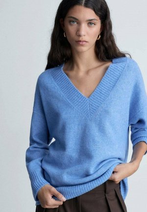Light blue oversized wool sweater with a deep V-neck and ribbed collar. Textured fabric and rolled sleeves, paired with dark brown pants.