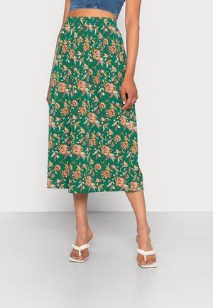 Vrouw draagt een groene midirok met een roze bloemenpatroon, een blauw cropped denim top en witte sandalen met hak.