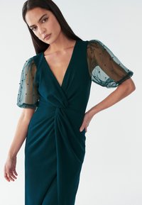 Teal jurk met een diepe V-hals, gegroepeerd twistdesign en doorschijnende puffmouwen versierd met zwarte kralen. Glad stof en elegante silhoutte.