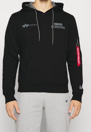 Mand iført sort Alpha Industries hættetrøje med grå snore, patches med teksten "Packers North Division" og rød mærkat på ærmet, kombineret med grå bukser.