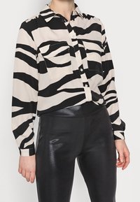 Svart och beige zebra-randig skjorta med knappar och långa ärmar, två bröstfickor och krage, i kombination med glänsande svarta leggings.