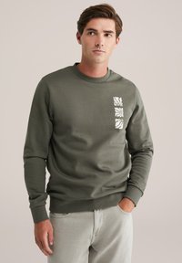 Olijfgroene sweatshirt met lange mouwen, ronde hals en een gestructureerd wit grafisch ontwerp op de borst. De stof lijkt zacht en comfortabel te zijn.