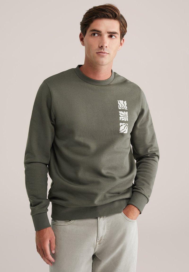 Olijfgroene sweatshirt met lange mouwen, ronde hals en een gestructureerd wit grafisch ontwerp op de borst. De stof lijkt zacht en comfortabel te zijn.