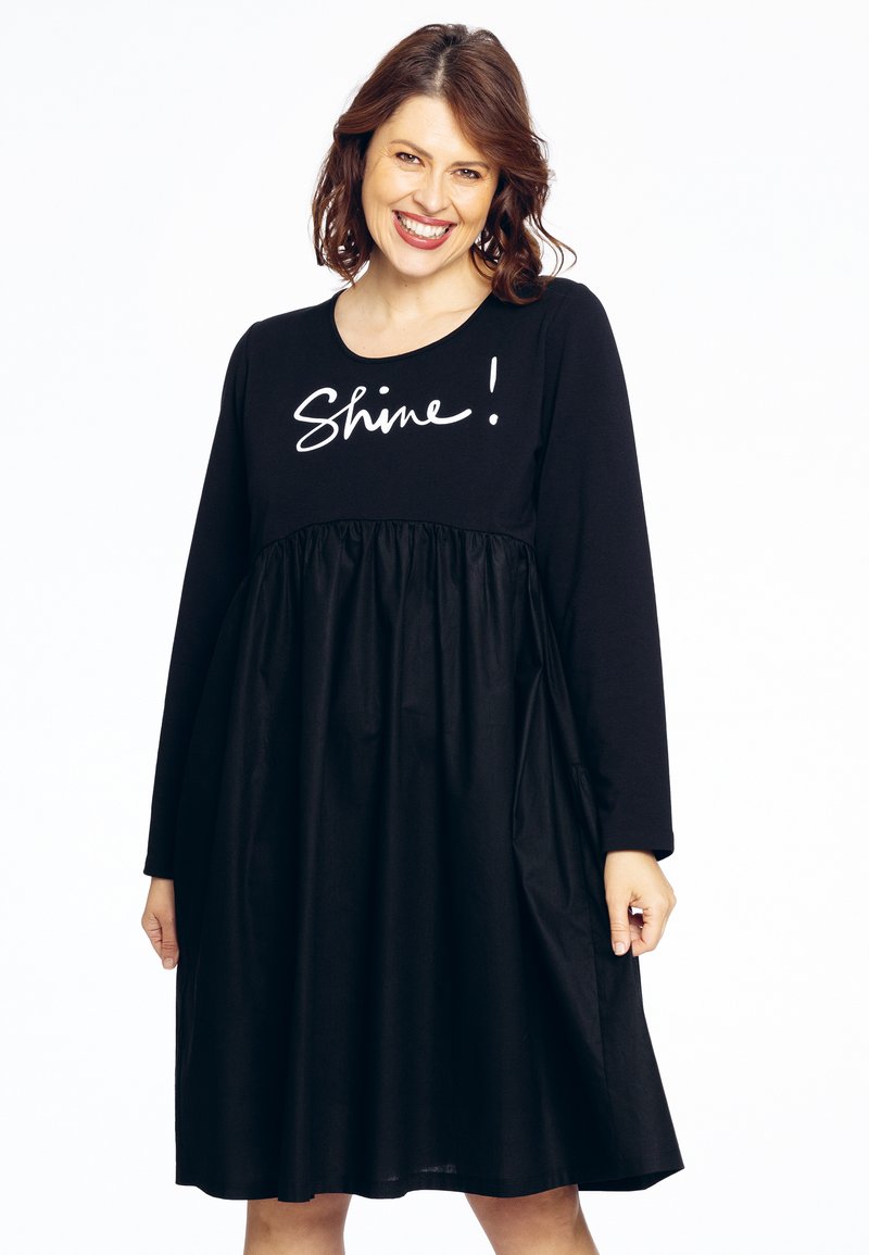 Sort kjole med lange ærmer, rynket krop og teksten "Shine!" i hvid kursiv. Glat stof med et afslappet design.