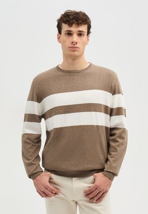 Giovane con capelli ricci che indossa un maglione marrone con due larghe strisce bianche orizzontali, in piedi con le mani vicino alle tasche.