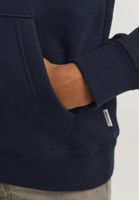 Sudadera azul marino con bolsillo tipo canguro, puños y dobladillo acanalados. Textura de tela suave con una pequeña etiqueta de marca en el lateral.