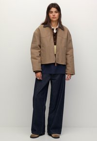 Ung kvinde iført en tan zip-up jakke, lagdelt strik, mørke brede jeans og beige sko, der står op ad en ensfarvet hvid baggrund.