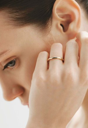 Primer plano de una persona tocándose la oreja con una mano que lleva un anillo de oro sencillo en el dedo medio sobre un fondo neutro.