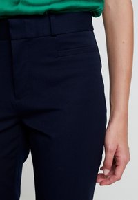 Pantaloni sartoriali blu navy con una texture di tessuto liscia, caratterizzati da una vestibilità slim, passanti per cintura e dettagli con tasche laterali.
