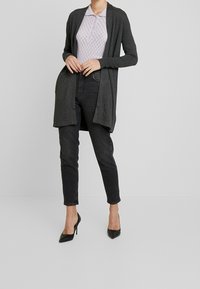 Langer, offen getragenes Cardigan aus dunkelgrauem Strick, über einem helllila Hemd mit Kragen und dunklen Jeans getragen, kombiniert mit schwarzen High Heels.