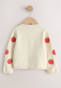 Pull de couleur crème suspendu sur un cintre en bois avec des motifs de fraises rouges sur les deux manches sur un fond uni.