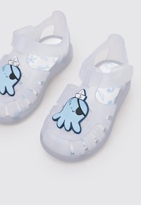 Sandali di plastica trasparente con grafica di un polpo blu sulla cinghia, con un sottopiede bianco e blu a fantasia e cinghia sul tallone regolabile.