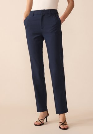 Mujer con pantalones rectos azul marino, de pie con las manos en los bolsillos, combinados con sandalias negras de tacón con tiras.