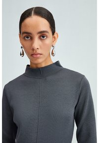 Top grigio a coste con colletto alto e maniche lunghe, caratterizzato da una cucitura centrale; modella che indossa orecchini a forma di goccia riflettenti.