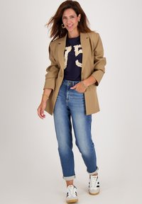 Khaki oversized blazer, marineblauwe shirt met textuurdetail "75", lichtblauwe opgerolde jeans en witte sneakers met zwarte strepen.