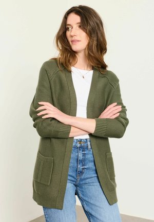 Woman with brown hair wearing a green cardigan, white shirt, and blue jeans stands with arms crossed looking to the side against a plain background.
