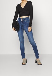 Svart långärmad omslagstopp, blå smala jeans med fem fickor och subtila slitningar, ihop med grå blockklack-mules och en svart fuskpälsväska.