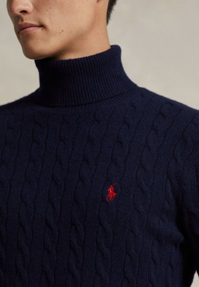 Uomo che indossa un maglione a collo alto lavorato a trecce blu navy con un piccolo logo ricamato rosso di cavallo e cavaliere sul petto.