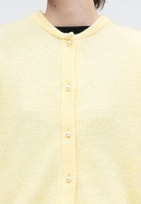 Κίτρινη κοντή ζακέτα με ριγέ υφή, διαθέτει στρογγυλή λαιμόκοψη και διακοσμητικά κουμπιά με πέρλες κατά μήκος του μπροστινού μέρους, παρουσιάζοντας ένα απαλό, υφασμένο πλέξιμο.