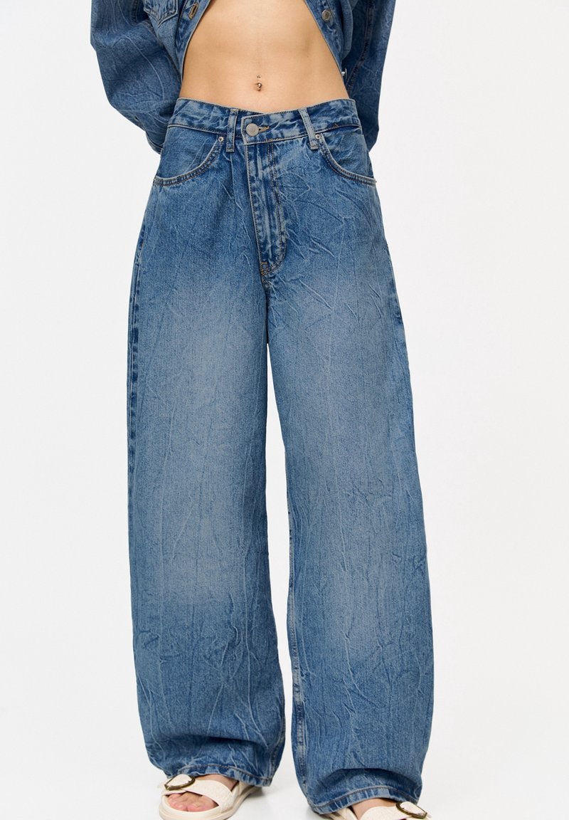 Jean denim bleu à jambes larges porté avec une veste en denim courte, dévoilant un ventre nu et des sandales blanches avec une boucle dorée.