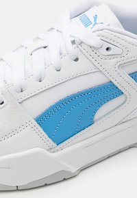 Baskets blanches avec un dessus en cuir lisse, présentant des accents bleu clair et des éléments de design superposés, complétées par des lacets et des coutures blancs.