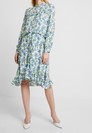 Kobieta w sukience w kwiaty z długim rękawem i falbaną na dole oraz białych szpilkach, stojąca na tle jednolitego koloru.