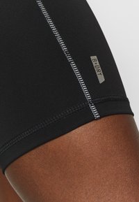 Sorte atletiske shorts lavet af strækbart stof, med reflekterende syninger og et sølvmærke med teksten "B:DRY" på siden.