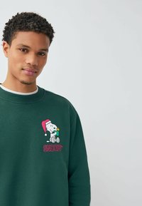 Zöld pulóver, kerek nyakkivágással, hímzett Snoopy mintával, aki Mikulás sapkát visel és egy virágot tart. A felirat alatt ez áll: "SNOOPY."