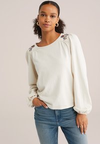 Blusa color crema, con texture, maniche a sbuffo e ricami floreali sulle spalle, abbinata a jeans blu. Tessuto morbido, vestibilità rilassata.