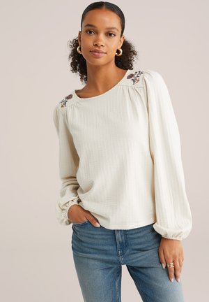 Cream-colored, textured blouse with puffed sleeves and floral embroidery on shoulders, paired with blue jeans. Soft fabric, relaxed fit.