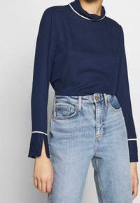 Granatowa bluzka z długim rękawem i wysokim kołnierzem, z białymi lamówkami i rozszerzanymi mankietami, w połączeniu z jasnoniebieskimi dżinsami z wysokim stanem.