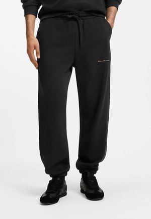 Uomo che indossa pantaloni jogger neri con polsini elastici e sneaker nere, in piedi con le mani in tasca su uno sfondo bianco semplice.
