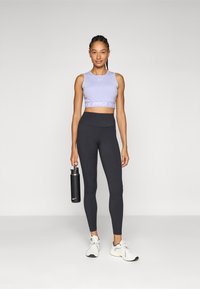 Paarse Nike Pro crop top met witte branding, gecombineerd met hoge zwarte leggings. Houdt een zwarte waterfles vast en draagt witte sneakers.